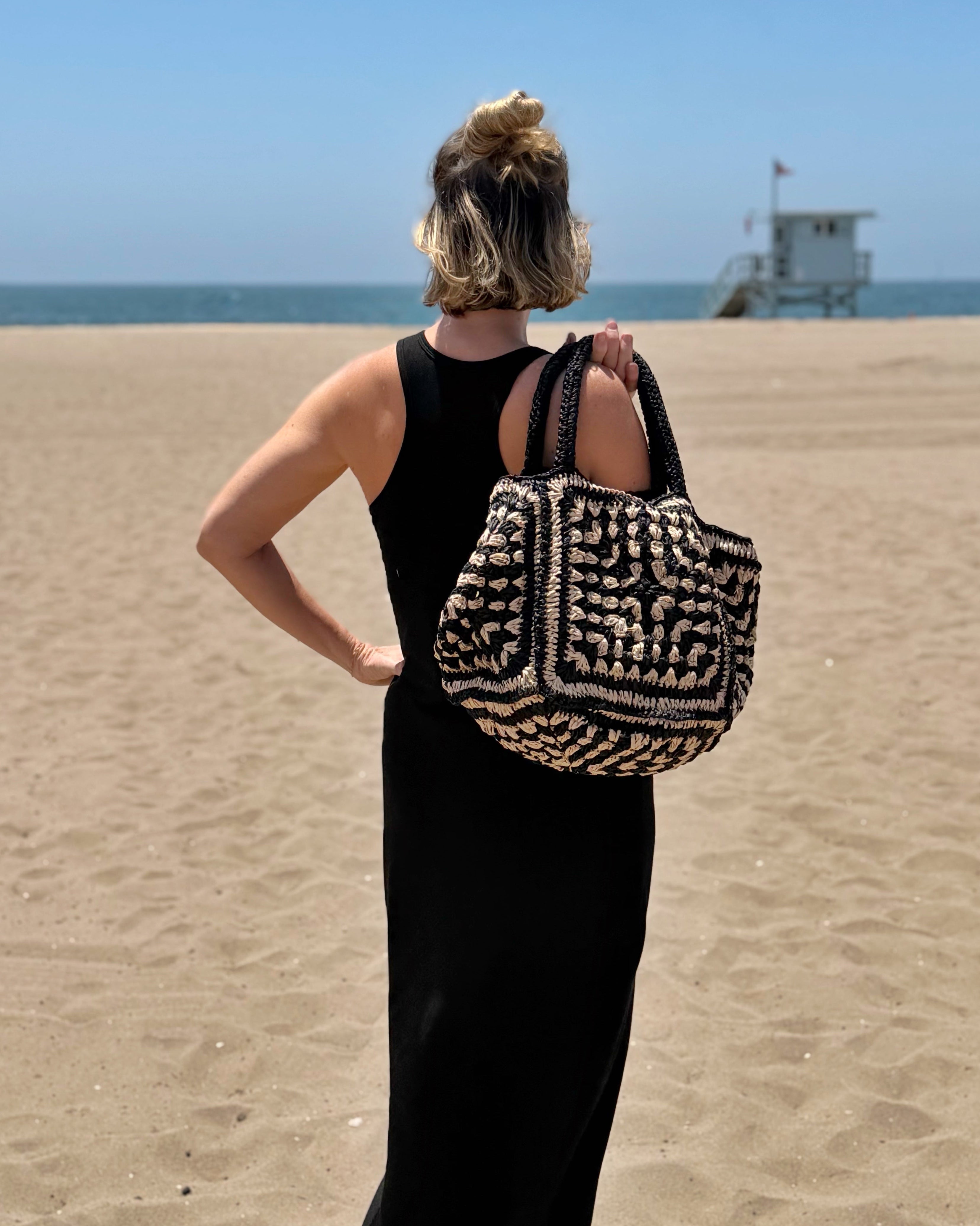 Person on a beach holding a patterned bag with a clear blue sky and ocean in the background. Mosaic  black and white beach tote 